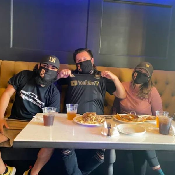 Three people wearing masks and hats, sitting at a table with food and drinks at R Bar.