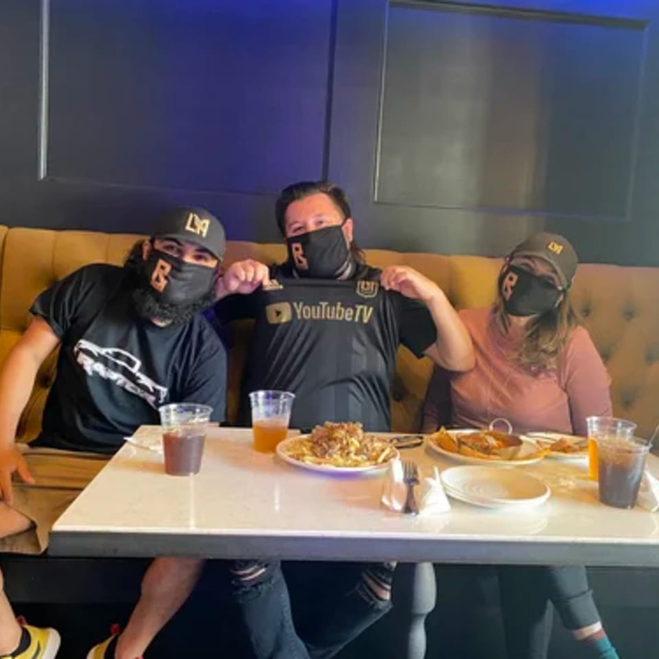 Three people wearing masks and hats, sitting at a table with food and drinks at R Bar.