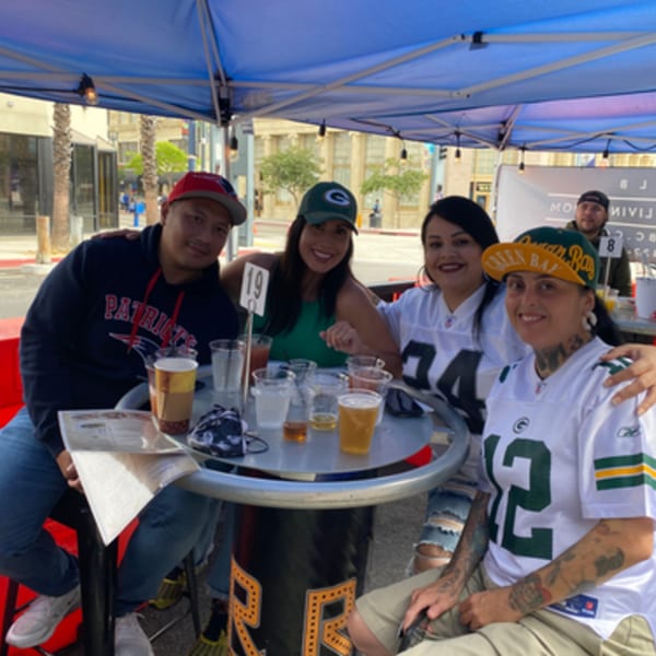 Four friends in sports jerseys and hats enjoy drinks at an outdoor R Bar table.