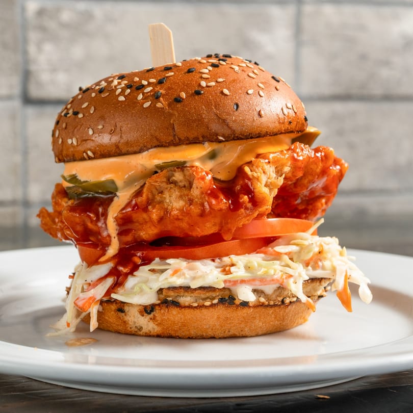 A crispy fried chicken sandwich with coleslaw, tomato, and sauce on a sesame bun.