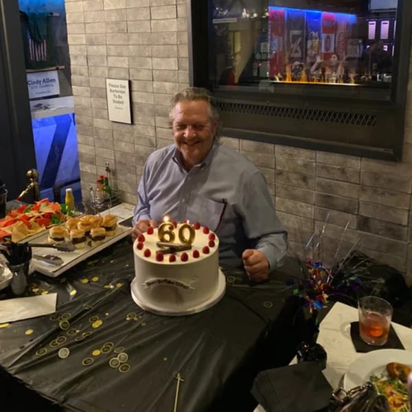 Man smiling behind a 60th birthday cake with raspberries, sliders, and chips at R Bar.