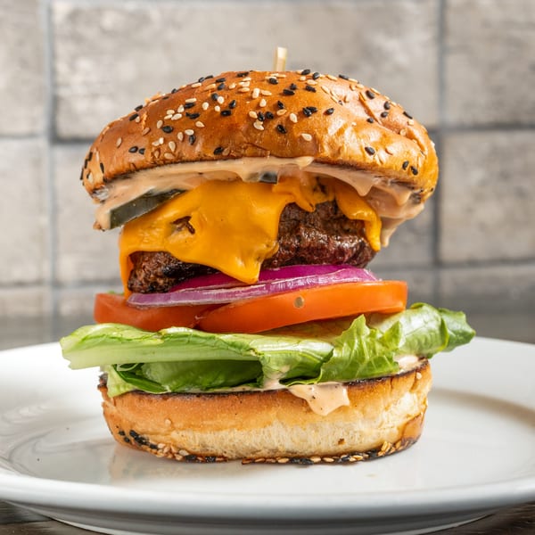 A tall R Bar burger with melted cheese, lettuce, tomato, onion, and sauce on a sesame bun.