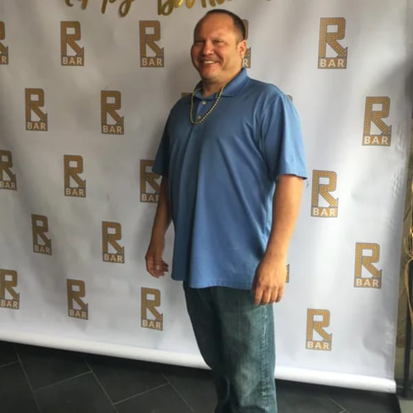 Man in a blue polo shirt and jeans smiling in front of an R Bar logo backdrop.