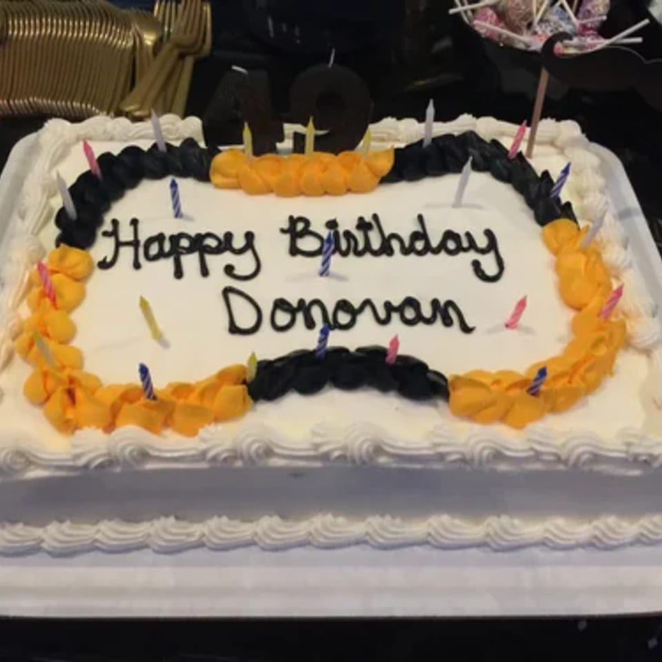 White birthday cake with "Happy Birthday Donovan" in black icing, surrounded by colorful candles.