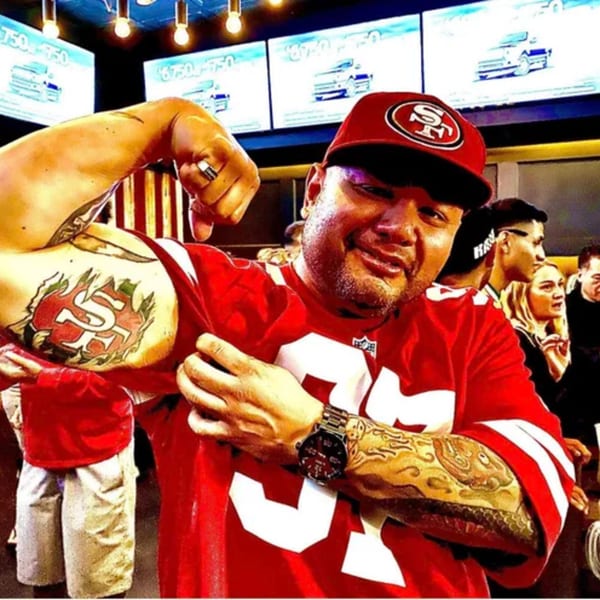 Man in a red 49ers jersey and cap flexes his bicep, revealing a 49ers tattoo at R Bar.