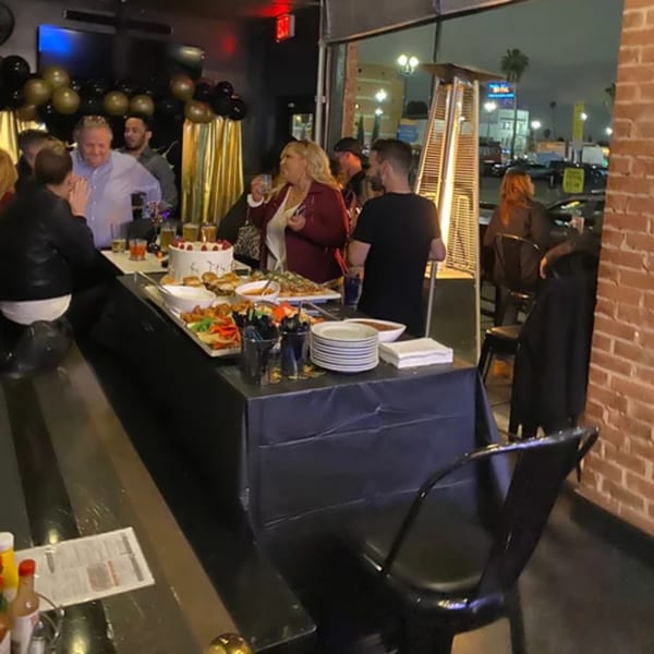 People gather around a buffet table with food and drinks at R Bar, decorated with balloons.
