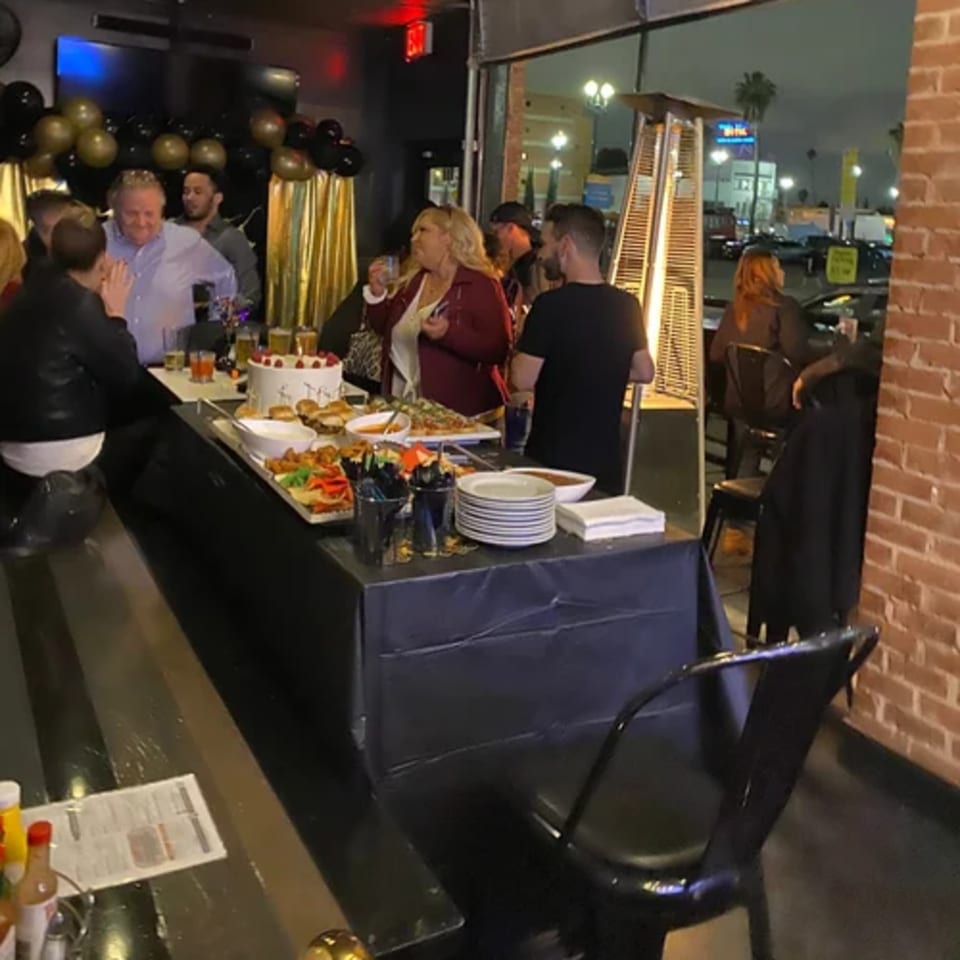 People gather around a buffet table with food and drinks at R Bar, decorated with balloons.