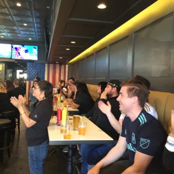 Patrons at R Bar clapping, watching a screen, with drinks and condiments on tables.