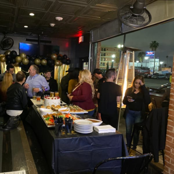 Guests mingle around a buffet table with food and drinks at R Bar, with city lights visible outside.