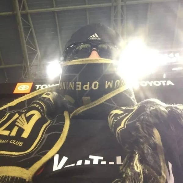 Person wearing a black Adidas hat, sunglasses, and a black and gold scarf with stadium lights.