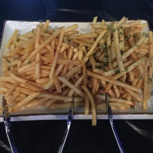 A white rectangular platter filled with golden french fries, some seasoned with herbs, and serving tongs.