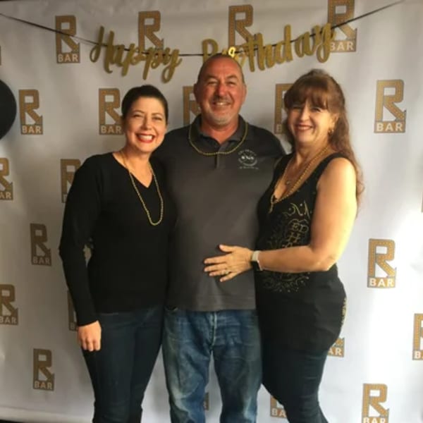 Three smiling people, two women and one man, posing in front of an R Bar branded backdrop.