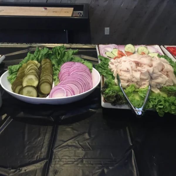 Buffet spread with sliced pickles, red onions, and deli meat on lettuce, with a shuffleboard table in the background.